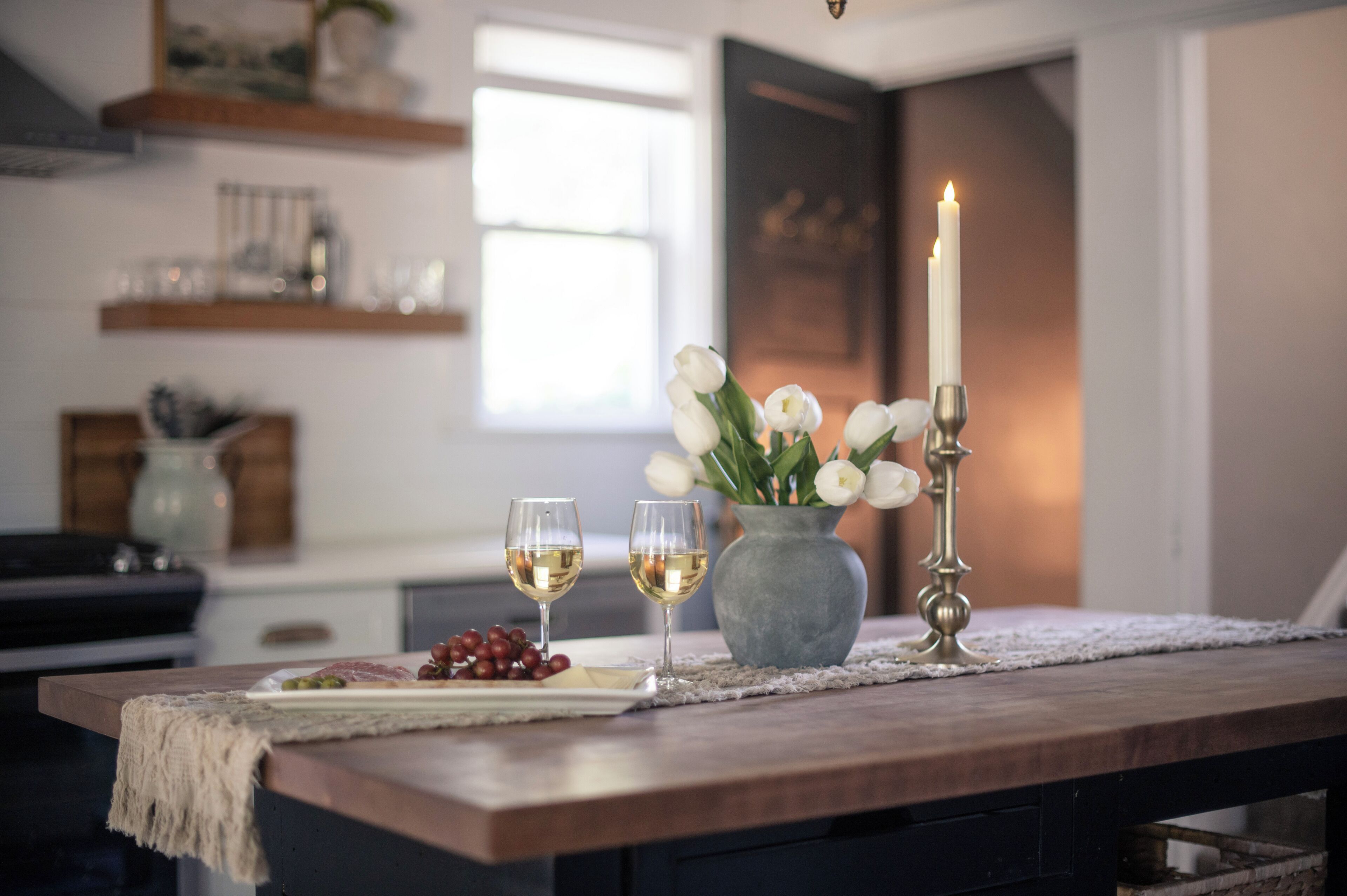Une table à manger avec deux verres de vin blanc, un vase de fleurs blanches et une bougie allumée.