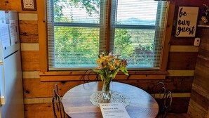Interior de uma cabana de madeira, com uma mesa redonda, um vaso de flores e uma placa de boas-vindas.