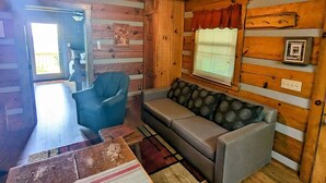 Interior acogedor de una cabaña con pared de madera, ventilador de techo, sofá, sillón y ventana con persianas.
