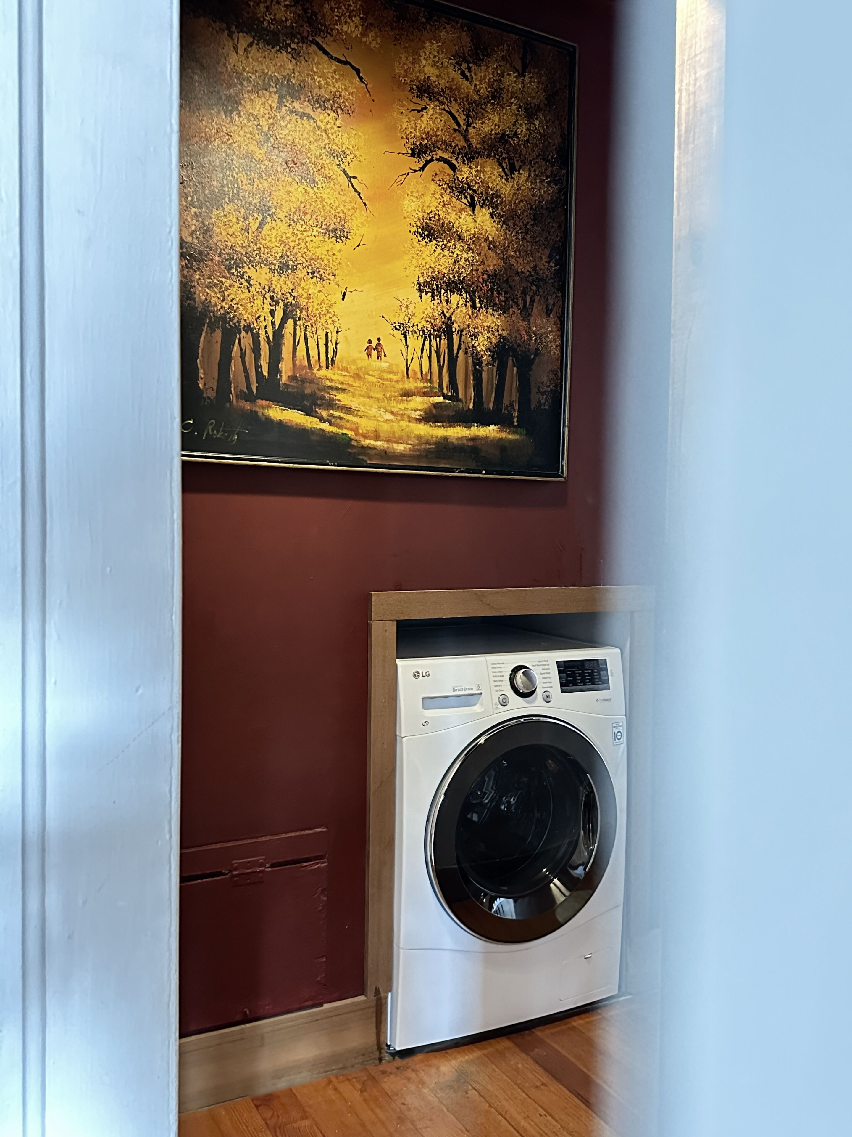 Ein Waschraum mit einer weißen Waschmaschine und einem Bild von einem von Bäumen gesäumten Weg darüber.