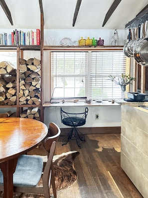 Une cuisine chaleureuse avec une table en bois, des chaises, un chien, une bibliothèque et une fenêtre avec des stores.