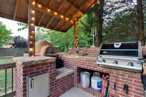 An outdoor barbecue area with a brick grill, a pizza oven, and string lights. - Fun in the sun - Outdoor fireplace, salt water pool, hot tub, sauna + much more (Greenbrier)