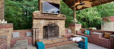 An outdoor patio with a stone fireplace, seating areas, and a television mounted above the fireplace.