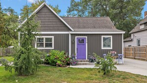 Una casa residenziale con facciata grigia, una porta d'ingresso viola e un piccolo giardino antistante con un albero e aiuole.