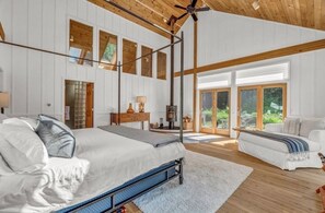 A spacious bedroom with a large bed, a wooden ceiling, and a view of the outdoors through large windows.