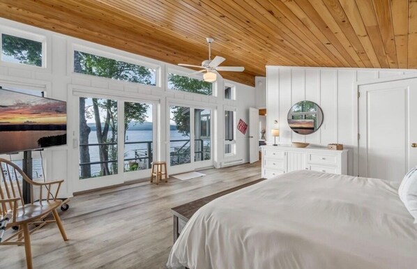 A bedroom with a large bed, a ceiling fan, a wooden floor, a desk with a chair, and a view of the outdoors through large windows.