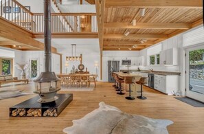 A spacious interior with a high ceiling, a central fireplace, a dining area, and a kitchen with large windows.