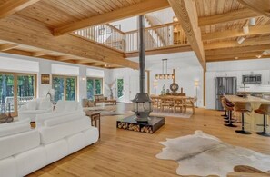 A spacious living area with a high ceiling, wooden beams, a central fireplace, and a dining area with a view of the outdoors.