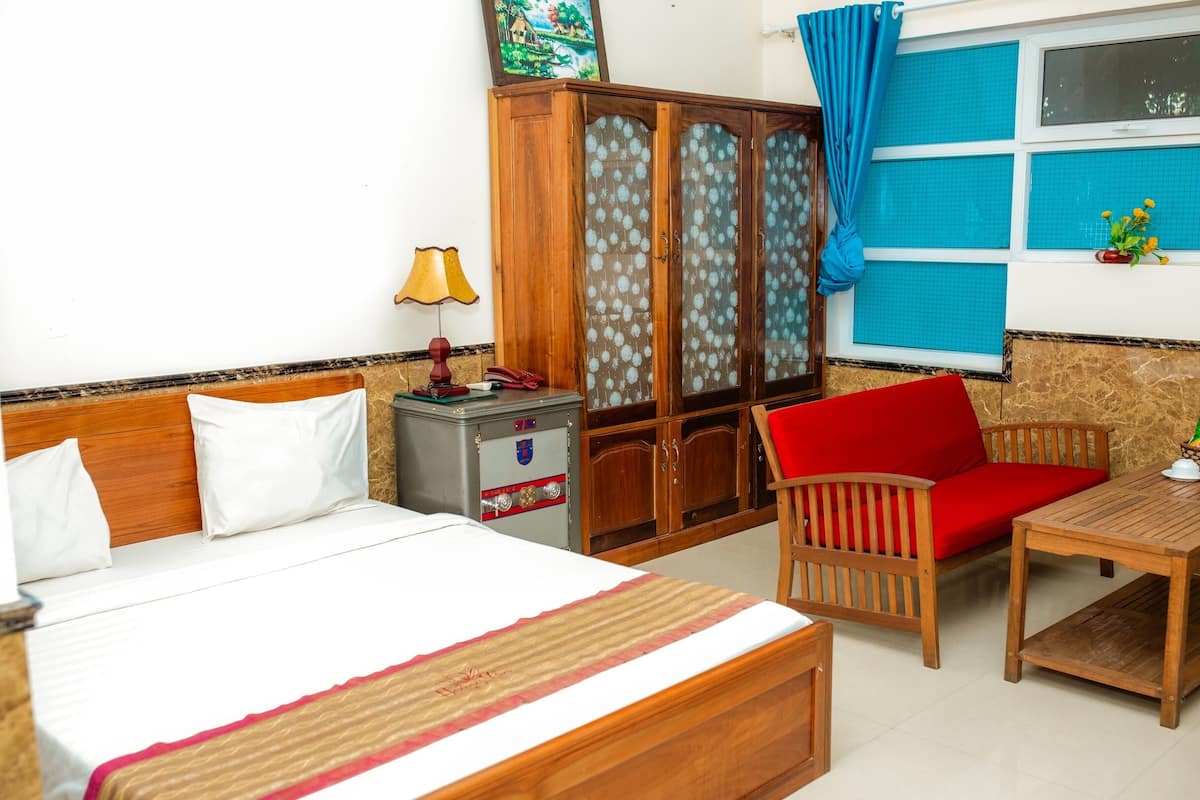 A hotel room with a bed, a wooden cabinet, a red chair, a small table, and a window with blue curtains.