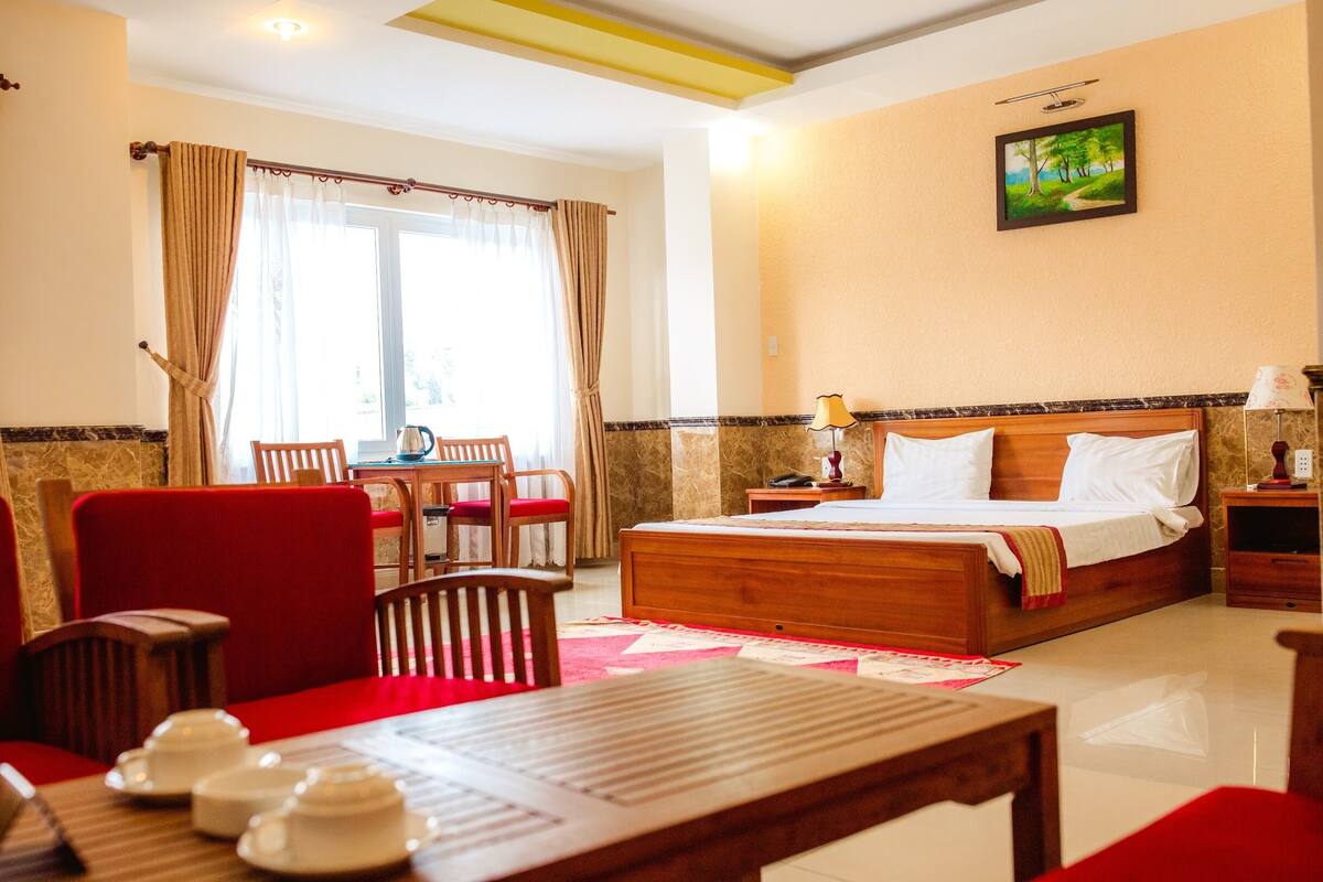 A hotel room with a bed, a dining table with chairs, a window with curtains, and a painting on the wall.