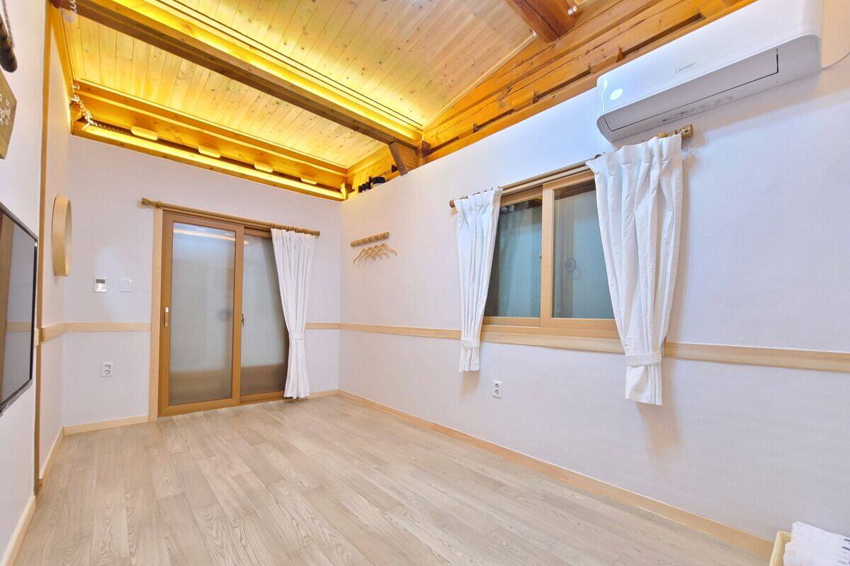 A spacious room with wooden flooring, a ceiling with exposed beams, and two windows with white curtains.