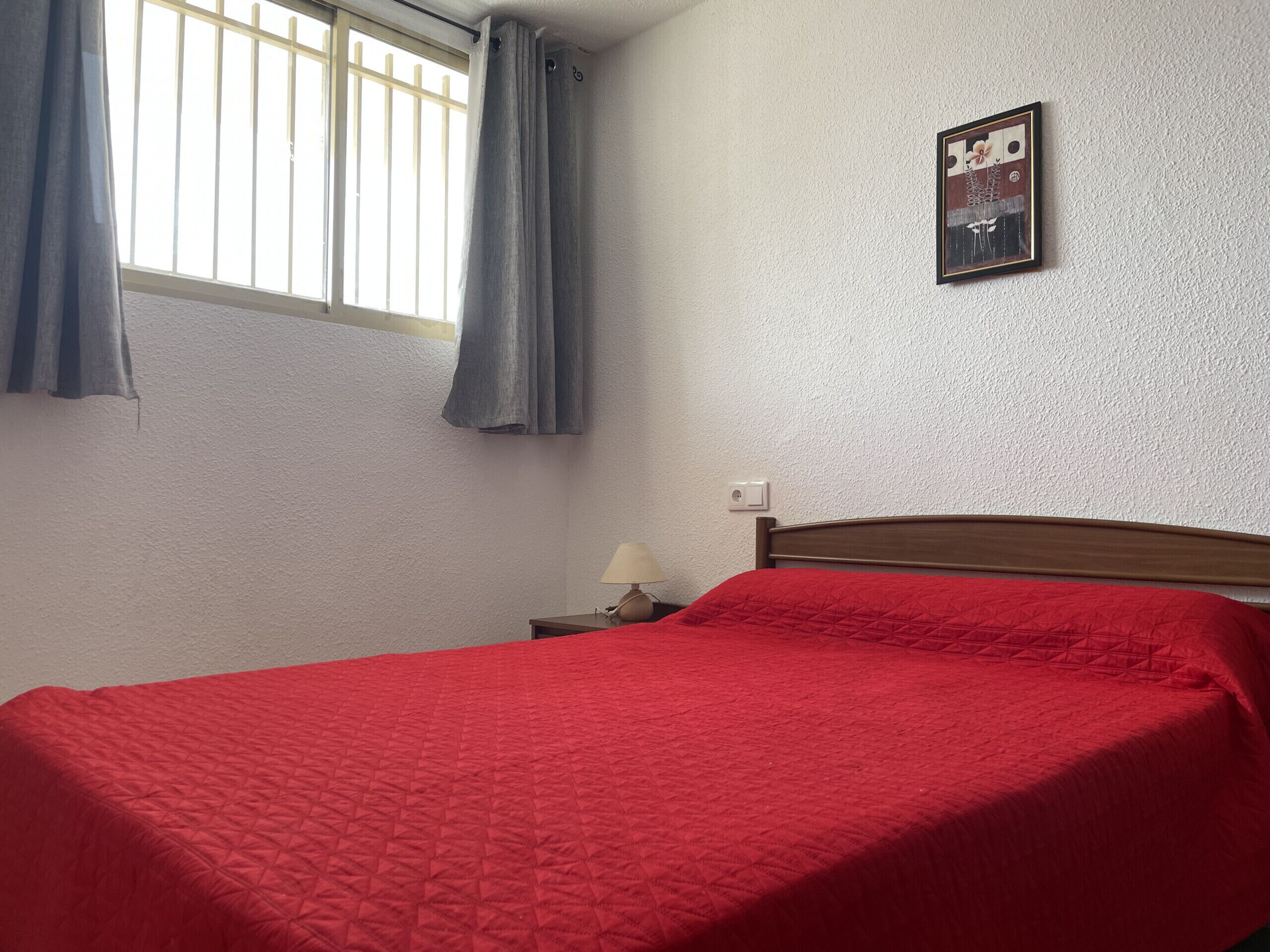 A room with a bed, a nightstand, a lamp, a window with curtains, and a wall-mounted picture.