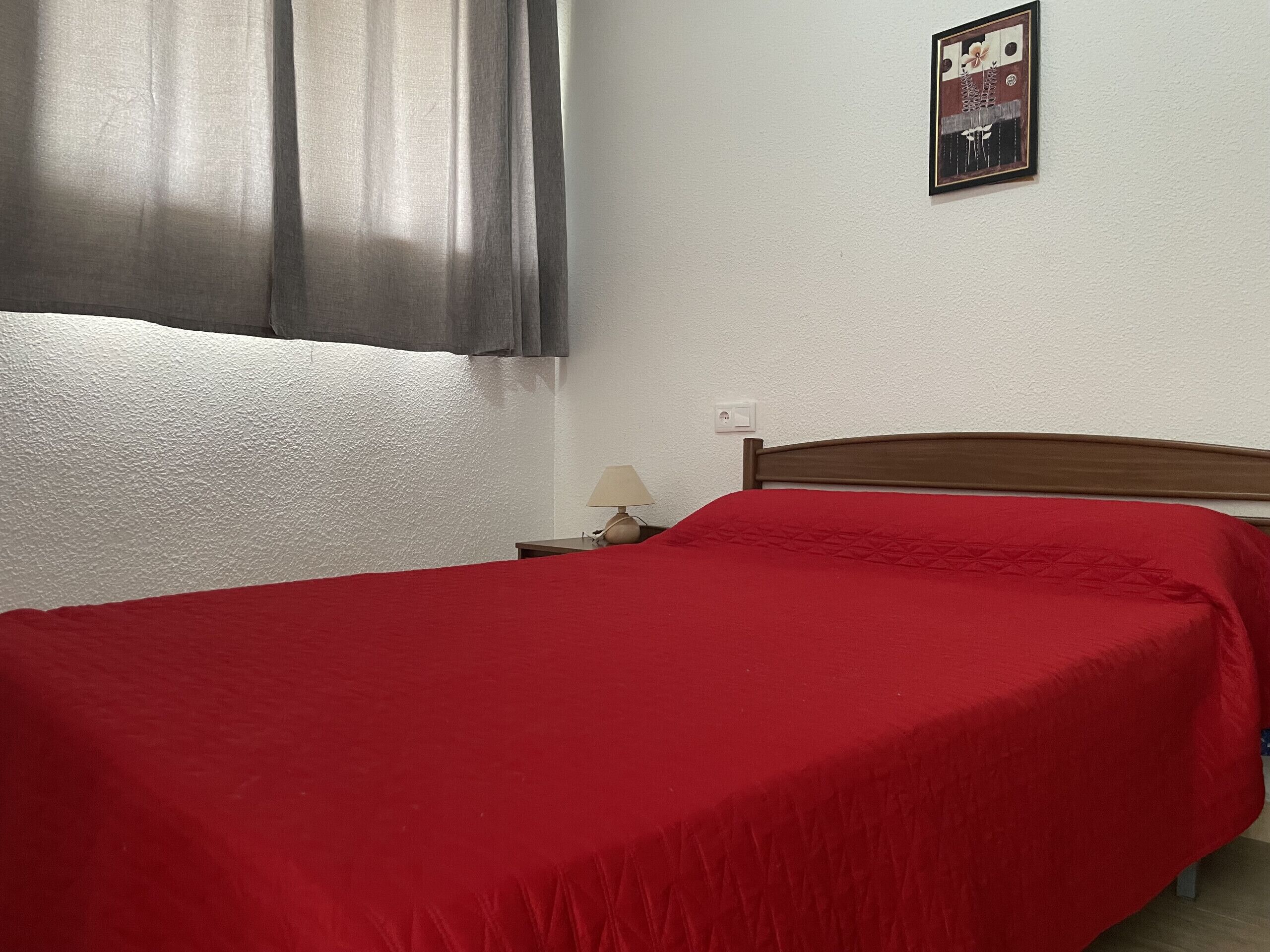 A single bed with a red bedspread in a room with white walls and a gray curtain.