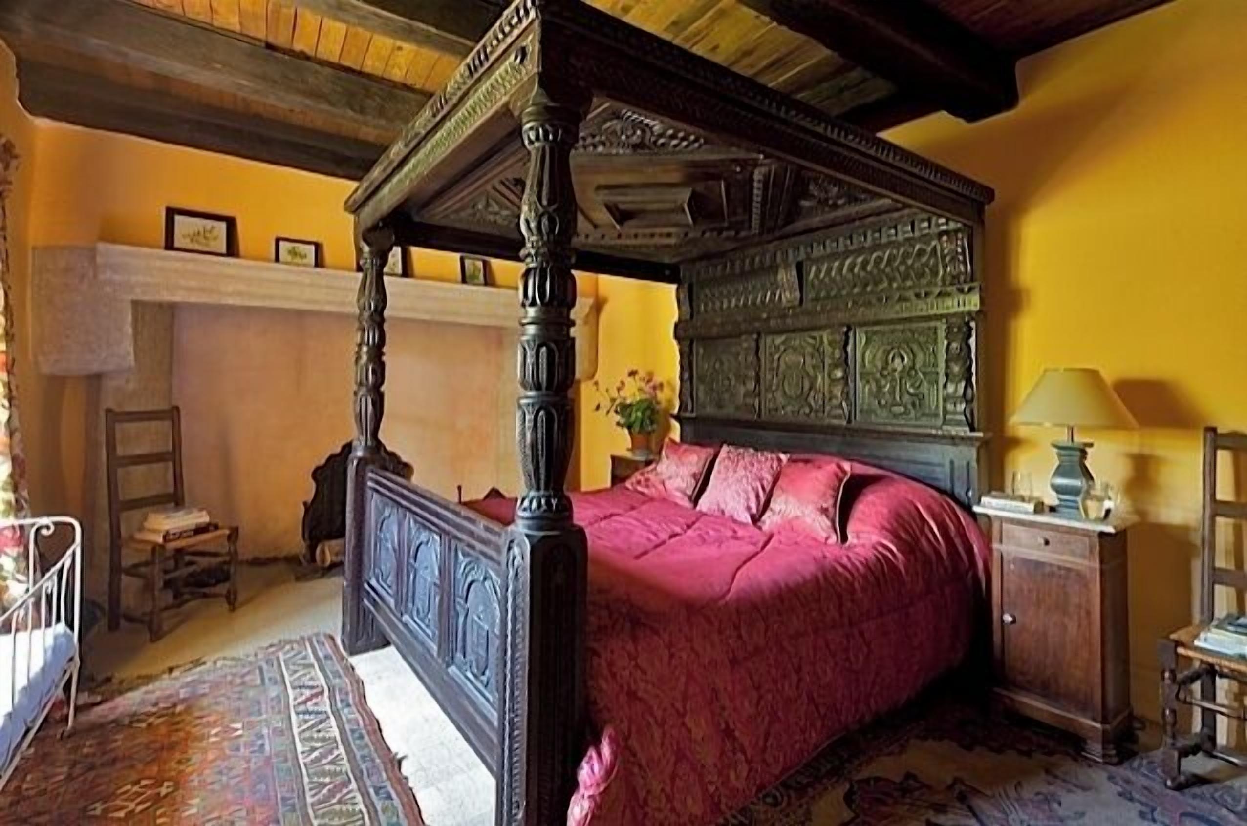A traditional four-poster bed with a red bedspread, a wooden nightstand with a lamp, and a decorative wall panel.