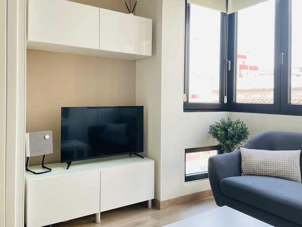A modern living room with a flat-screen TV on a white cabinet, a grey sofa, and a window with blinds.