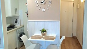 Une petite cuisine avec une table blanche, deux chaises, une horloge, un lave-linge et un placard.