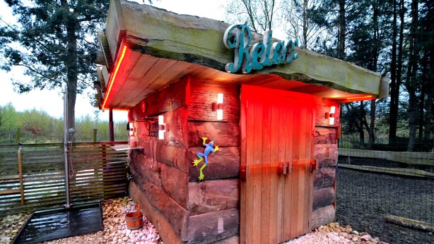 Una cabaña de madera rústica con una puerta roja, un cartel sobre la puerta y una ventana pequeña.