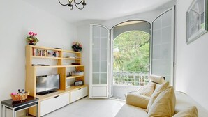 A living room with a sofa, a TV on a wooden stand, a bookshelf, and a sliding glass door leading to a balcony with greenery outside. - A beautiful apartment tastefully decorated ... (Nice)