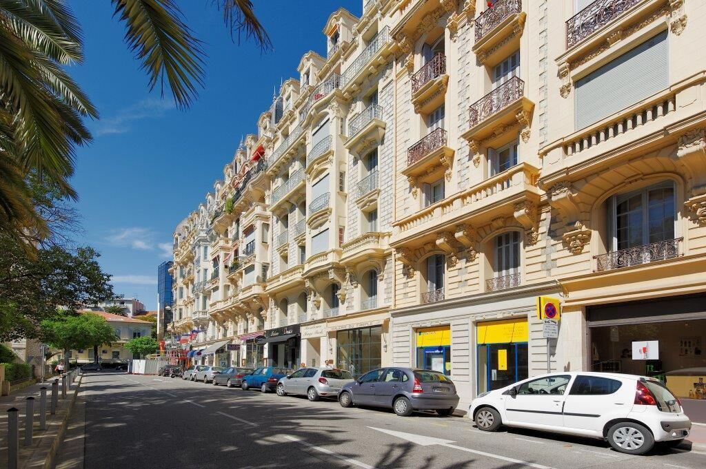 A row of multi-story buildings with balconies, parked cars, and palm trees.