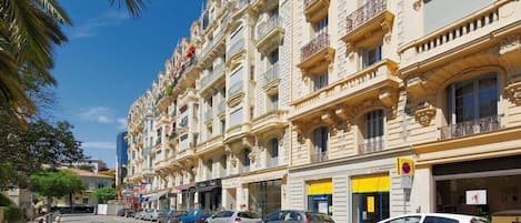 A row of multi-story buildings with balconies, parked cars, and palm trees.