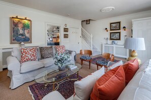 A living room with a white sofa, a glass coffee table, and patterned cushions. There is a staircase leading to another room, and a door with a white frame.