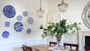 Uma sala de jantar com uma mesa de madeira, cadeiras, um vaso com flores e pratos com padrão azul e branco na parede.