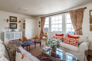 A well-lit living room with a white sofa, patterned cushions, a glass coffee table, and a decorative vase. There are two chairs, a small side table, and a white cabinet with framed pictures.
