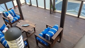 A covered outdoor seating area with wooden furniture and cushions, a table with a fish-shaped object, and large windows offering views of the outside.