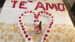 A bed with a heart-shaped arrangement of rose petals spelling "TE AMO" and a bottle of champagne.