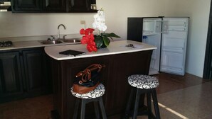 A kitchen with dark cabinets, a white countertop, a sink, a refrigerator, and two stools with patterned cushions.