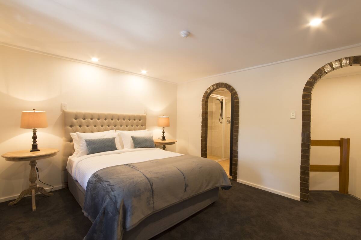 Un dormitorio con una cama grande, dos mesitas de noche con lámparas, cabina de ducha de vidrio y un arco de ladrillo.