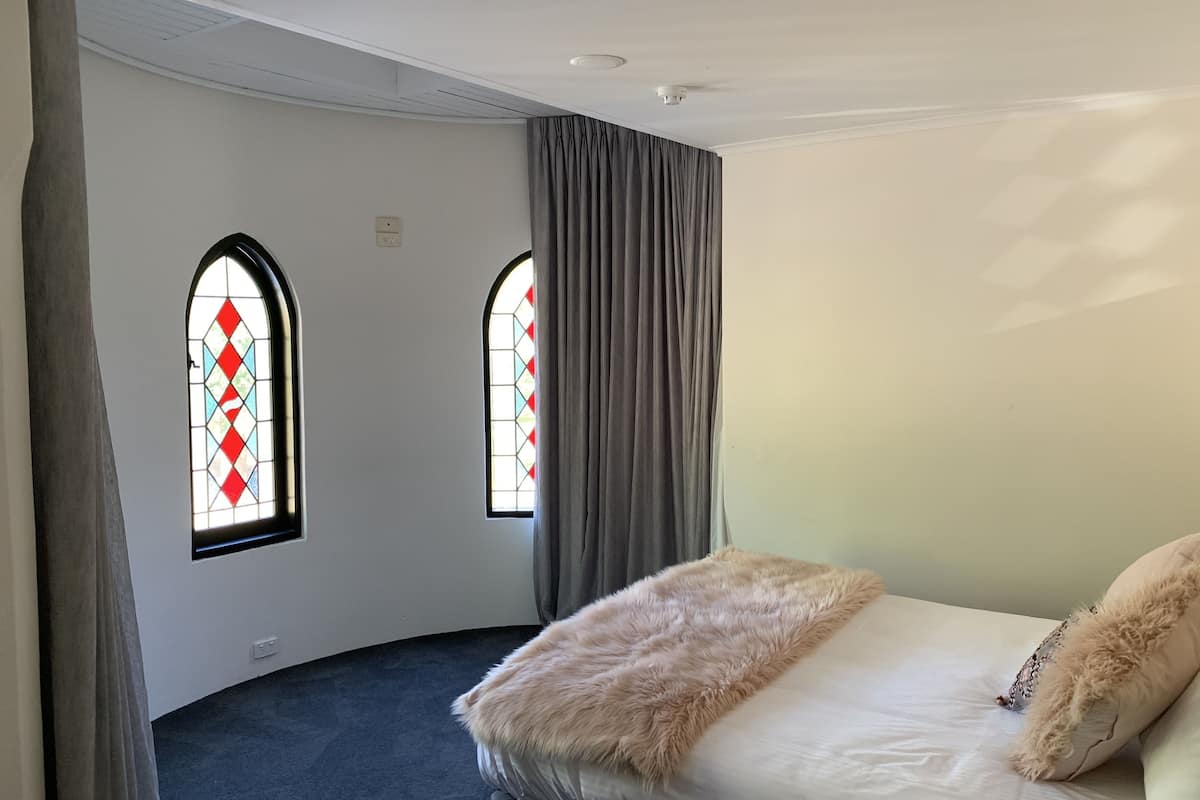 Un dormitorio con una cama, dos ventanas con cortinas, una lámpara en el techo y suelo cubierto de alfombra.