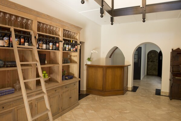 Uma adega com prateleiras de madeira, garrafas e copos de vinho. Há uma escada, uma pequena mesa com um vaso e uma parede de pedra ao fundo.