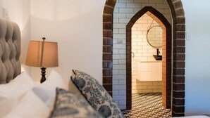 A bedroom with a bed, a lamp, patterned pillows, and an arched doorway leading to a bathroom with a tiled floor. - The Manor Basket Range (Basket Range)