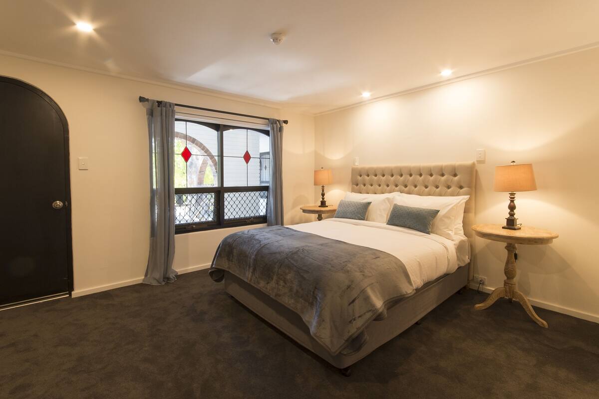 Un dormitorio con una cama, dos mesitas de noche con lámparas, una ventana con cortinas y una puerta.