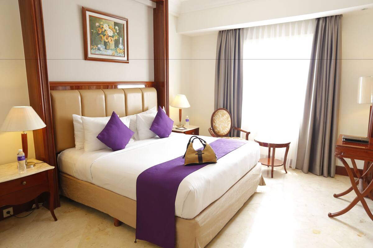 Una habitación de hotel con una cama grande, una mesita de noche con lámpara, una silla, un escritorio y una ventana con cortinas.