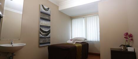 Habitación de hotel con cama, mesita de noche con un jarrón de flores, ventana con persianas, lavamanos empotrado en la pared y un cuadro colgado.