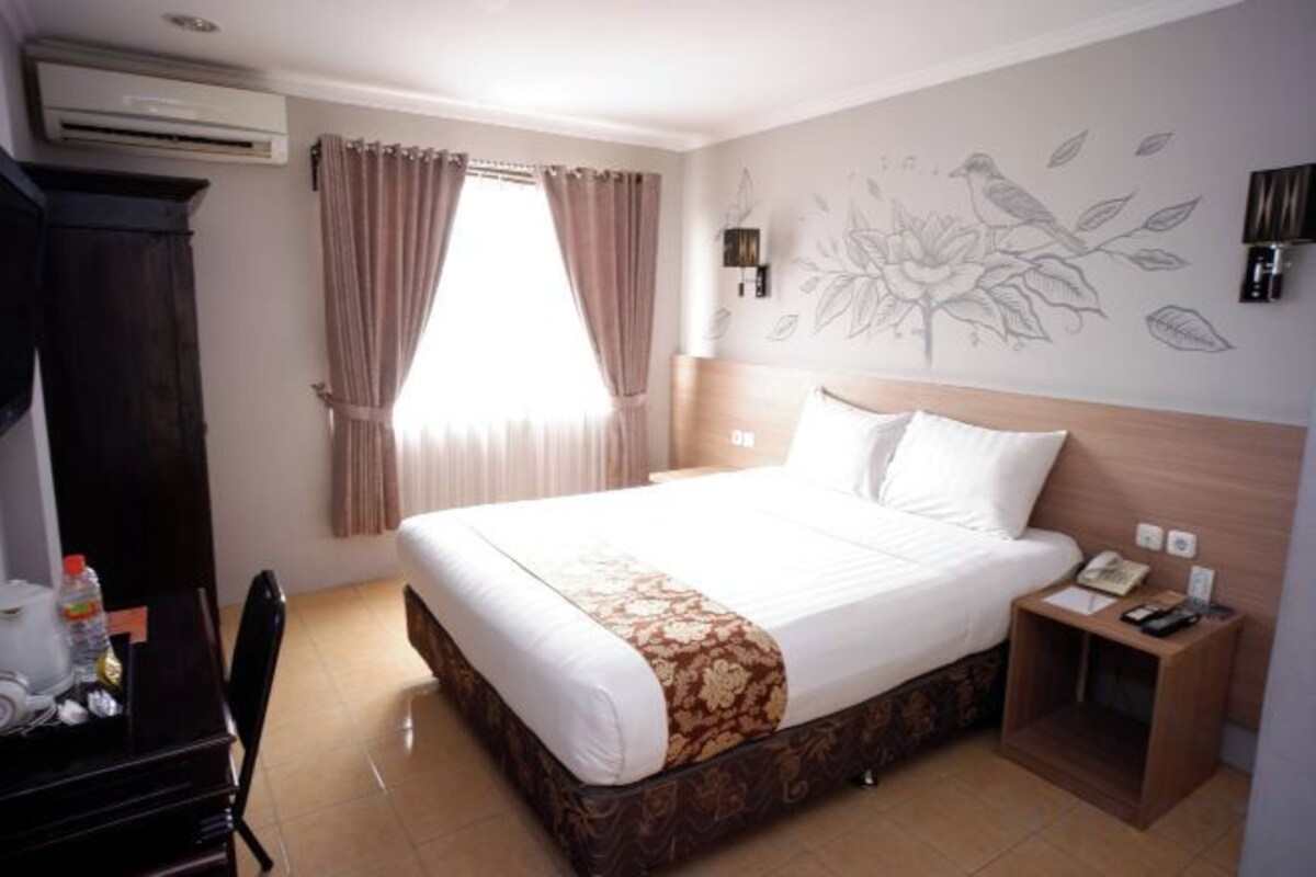 A hotel room with a large bed, a desk with a chair, a bedside table with a phone, and a wall with a floral design.