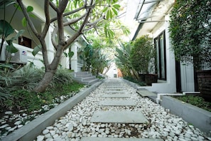 Un sendero bordeado de guijarros blancos y un árbol a la izquierda, que conduce a un edificio de fachada blanca y una escalera.