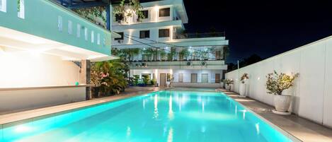 A swimming pool with a clear blue water surface, surrounded by a white wall and a building with multiple balconies.