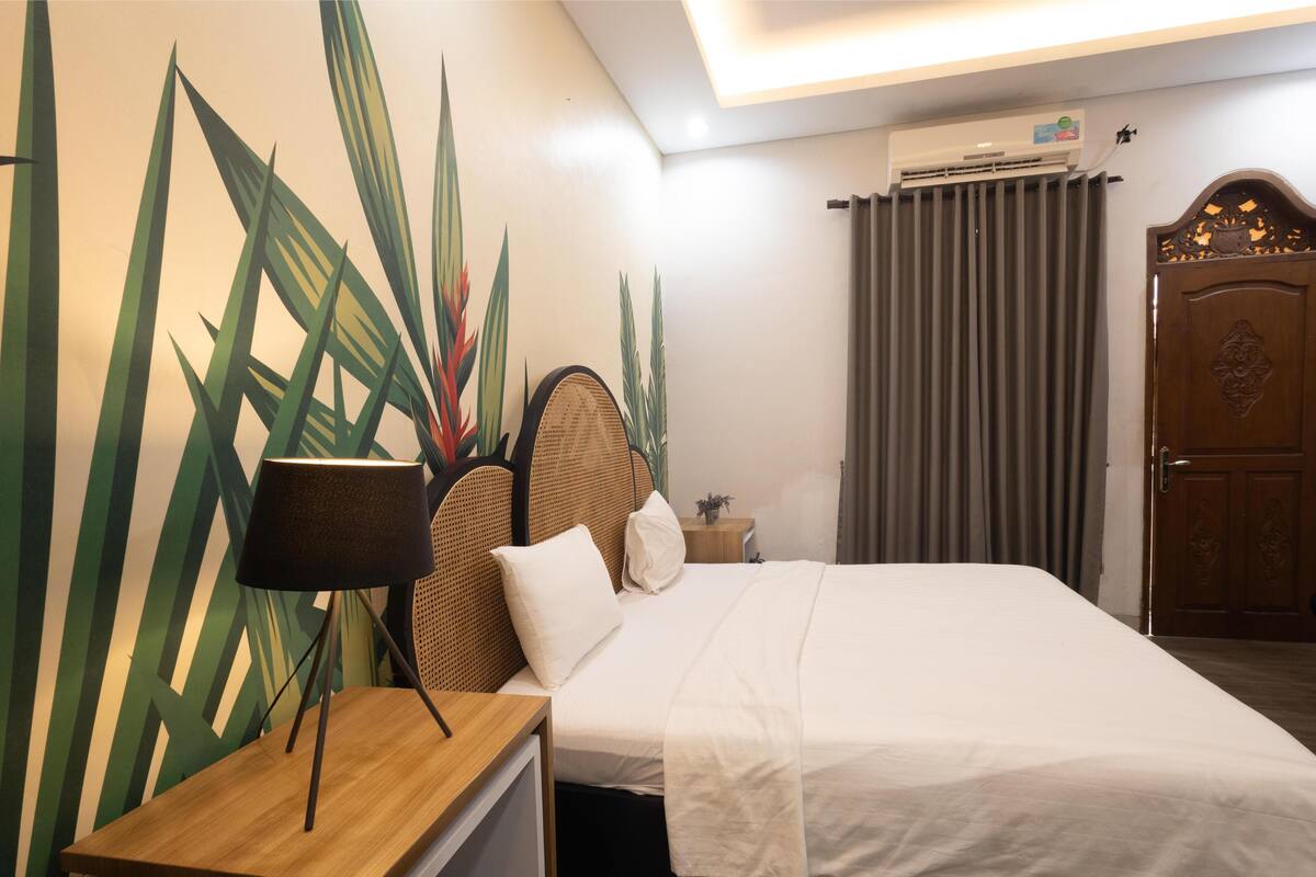 A hotel room with a bed, a bedside table with a lamp, a wall-mounted air conditioner, a curtain, and a wooden door with a decorative panel.
