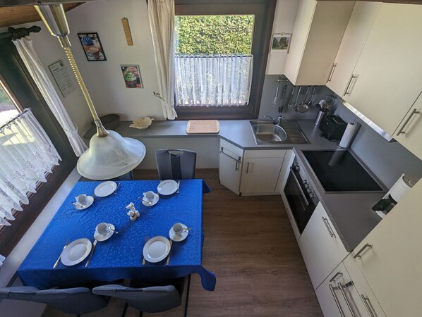 A compact kitchen with a dining area, featuring a blue tablecloth, white cabinets, and a window with a view of greenery.