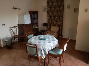 A dining area with a round table covered by a checkered tablecloth, surrounded by wooden chairs with cushioned seats. A refrigerator is visible in the background, and a bookshelf is against the wall.
