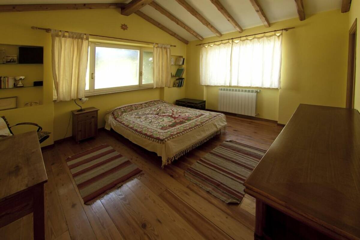 A bedroom with a large bed, wooden floor, two windows with curtains, a radiator, and a wooden table.
