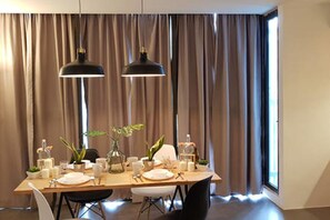 A dining area with a table set for four, two pendant lights, and a view through large windows with curtains. - Duplex Loft Empire Damansara (Petaling Jaya)