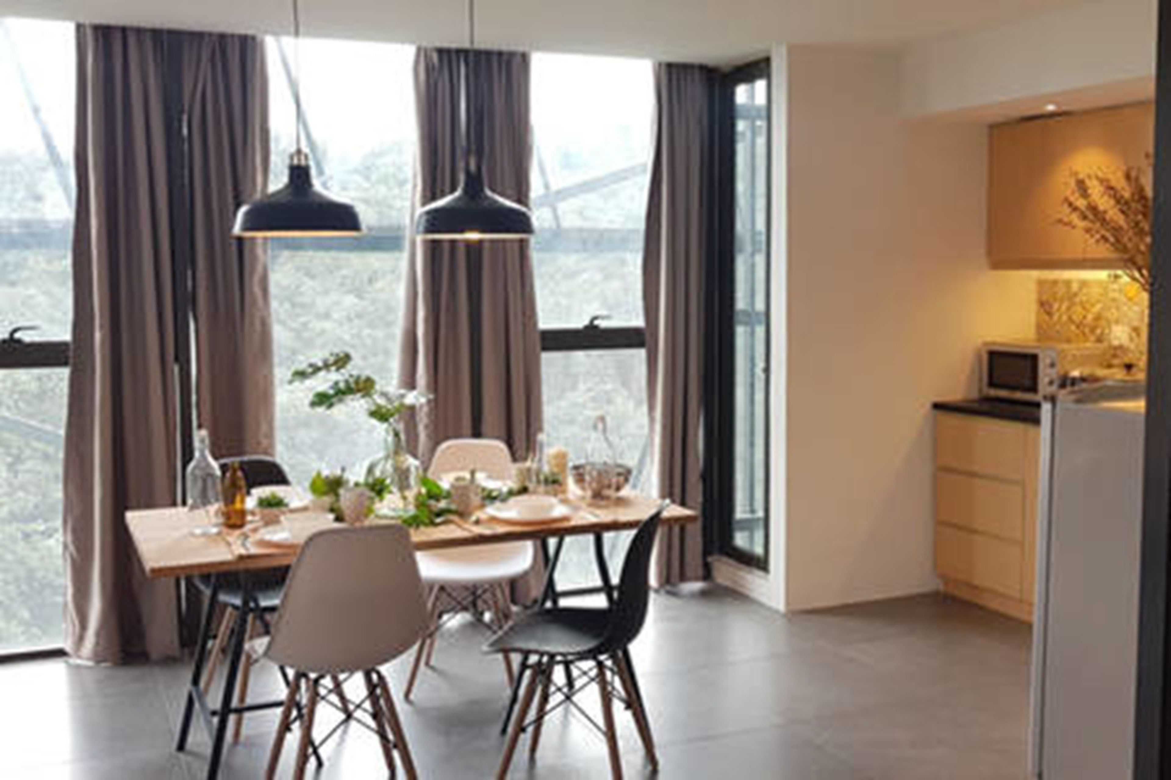 A dining area with a table set for four, two pendant lights, and a view through large windows.