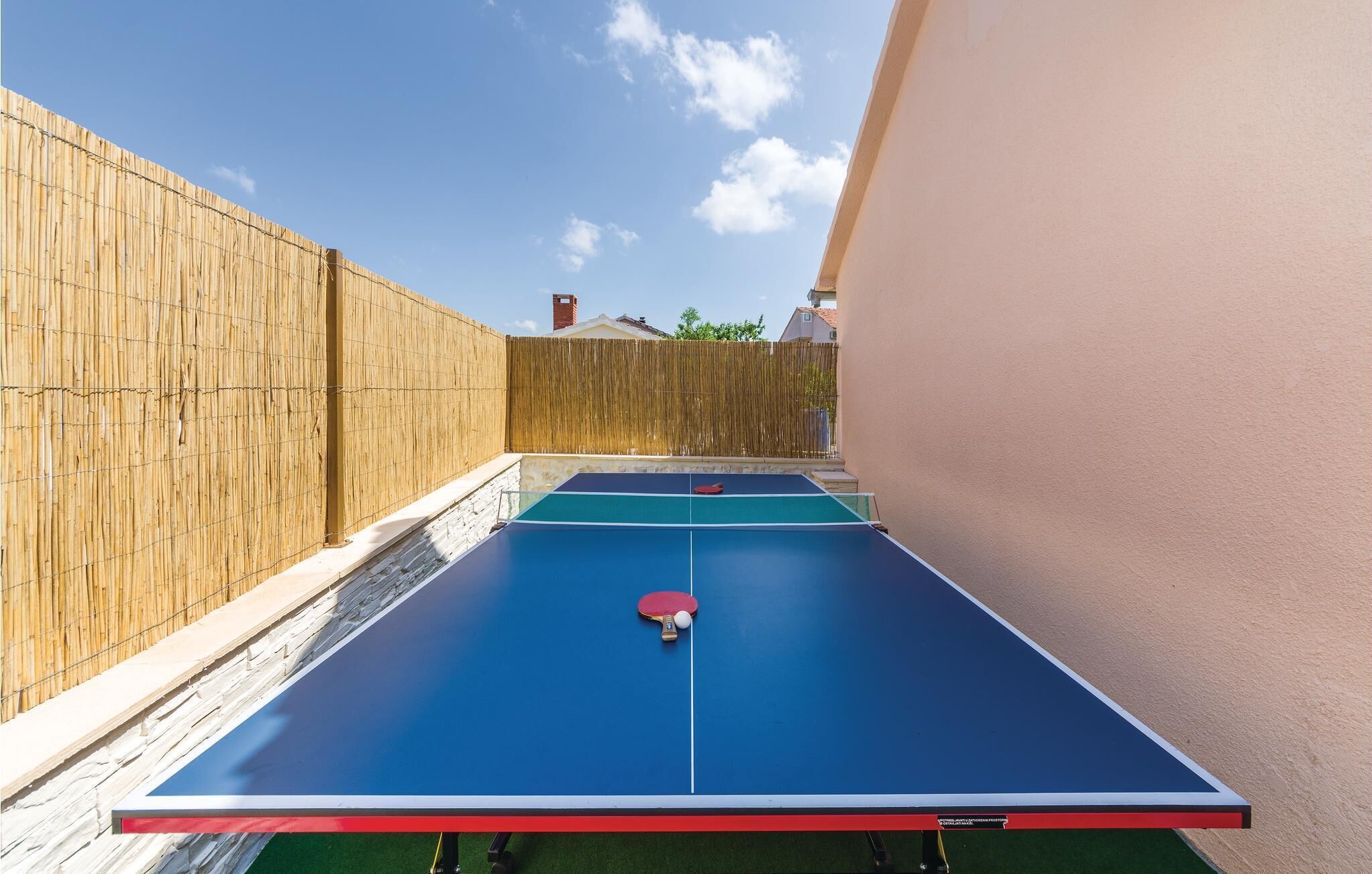 An outdoor ping pong table with a net in the middle, surrounded by a bamboo fence and a beige wall.