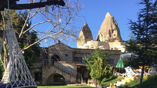 A traditional stone building with a large central tower and two smaller towers, surrounded by a well-maintained garden with a reflective pool in the foreground. - Kufe Hotel (Nevsehir)