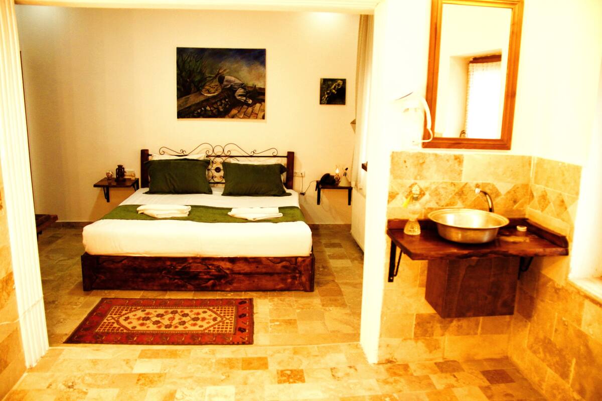 A bedroom with a bed, a nightstand, a painting, a mirror, and a bathroom with a sink and a bowl.