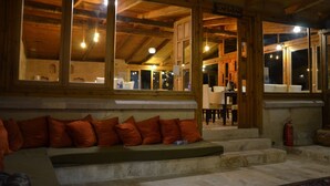 A wooden cabin with a stone bench and orange cushions, a cafe-bar area, and large windows. - Kufe Hotel (Nevsehir)